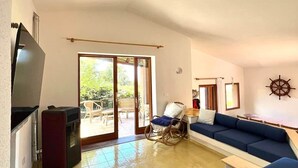 A spacious living room with a large blue sofa set, a flat-screen TV, and a sliding glass door leading to an outdoor area. - Domus Ninni - Charm of Gallura (Costa Paradiso)