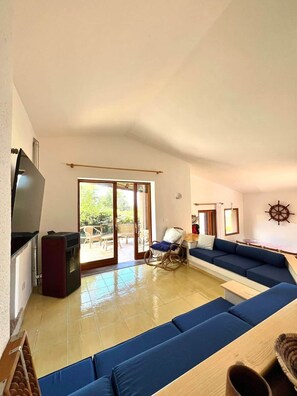 A spacious living room with a large blue sofa set, a flat-screen TV, and a sliding glass door leading to an outdoor area. - Domus Ninni - Charm of Gallura (Costa Paradiso)