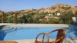 A swimming pool with a wicker chair in the foreground, a metal ladder, and a view of a hillside with buildings. - Domus Ninni - Charm of Gallura (Costa Paradiso)