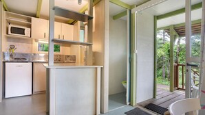A compact kitchen with a microwave, stove, and cabinets, a small seating area with a chair, and a view of greenery through a glass door.