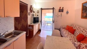 Un espacio habitable compacto con cocina, comedor y un sofá con cojines rojos. Un televisor está montado en la pared y se aprecia vista al exterior a través de una puerta abierta.