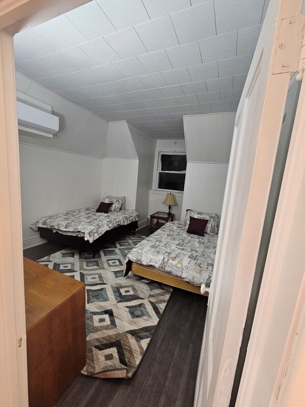 A small, compact room with two beds, a wooden bench, a patterned rug, and a window.