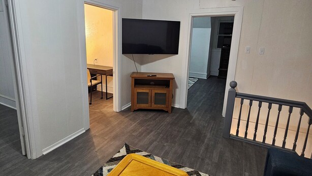 Ein Wohnzimmer mit einem an der Wand montierten Flachbildfernseher, einem Holztisch, einem dunkelgrauen Sofa und einem gemusterten Teppich.
