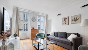 Un salon moderne avec un canapé, une table basse en verre, un meuble en bois et un fauteuil vert. Il y a deux fenêtres avec des rideaux, une horloge murale et deux reproductions botaniques encadrées.