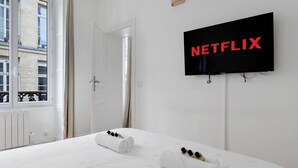 Une chambre d’hôtel avec un lit, un téléviseur fixé au mur diffusant du contenu « NETFLIX », et une fenêtre donnant sur des immeubles.