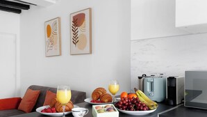 Eine moderne Küche mit Marmorfliesenspiegel, einer Kaffeemaschine und einem Toaster. An den Wänden hängen zwei gerahmte Kunstwerke, ein Sofa mit orangefarbenen Kissen und ein Frühstückstisch mit Croissants, Obst und einem Glas Orangensaft.
