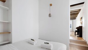 Ein schlicht eingerichtetes Schlafzimmer mit einem Bett, zwei Handtüchern, einer Pendelleuchte und einem Klavier im angrenzenden Raum.
