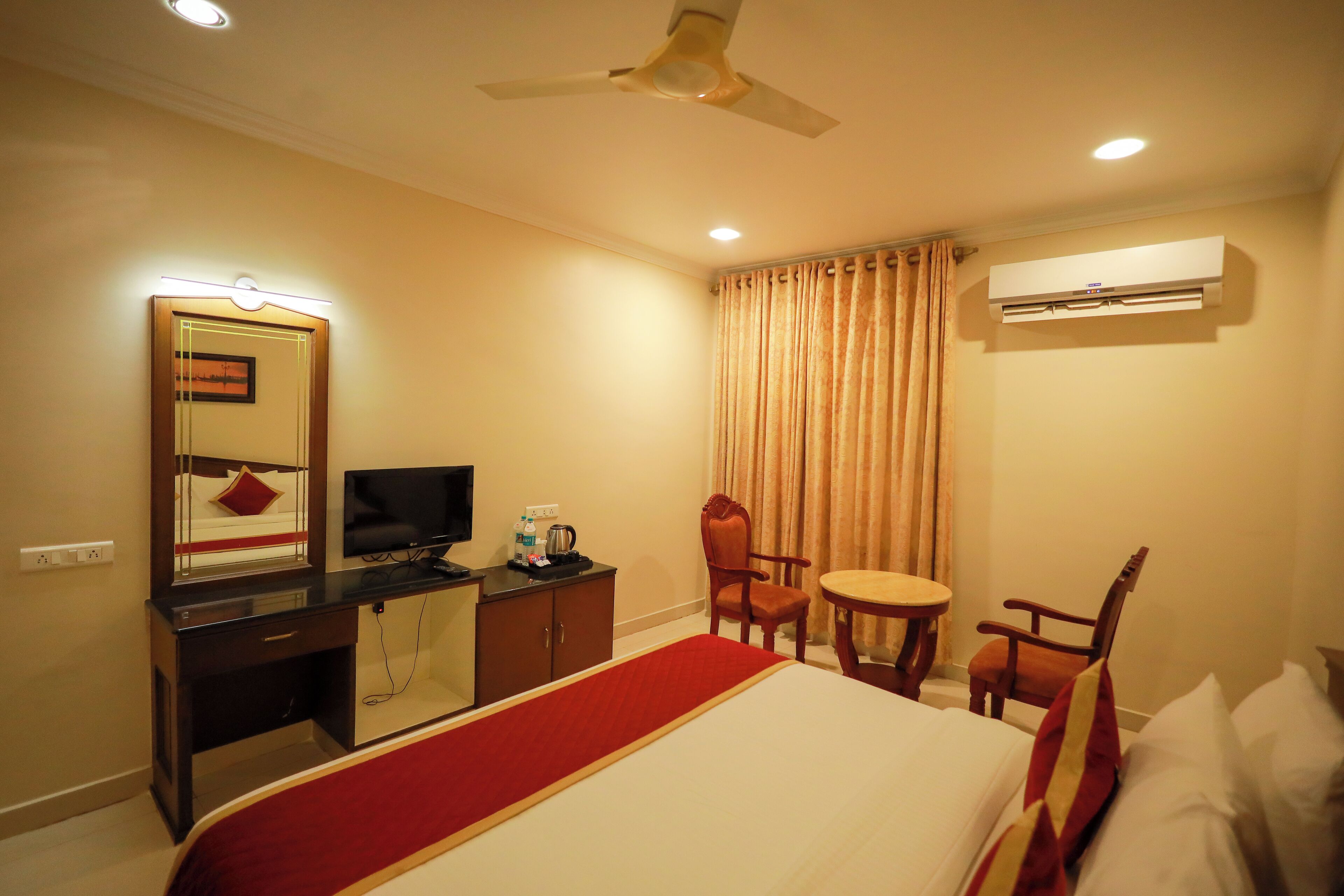 A hotel room with a bed, a desk with a TV, a chair, a small table, and an air conditioning unit.