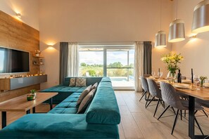 Ein modernes Wohnzimmer mit einem türkisfarbenen Sofa, einem Esstisch für sechs Personen und einem großen, an der Wand befestigten Fernseher.