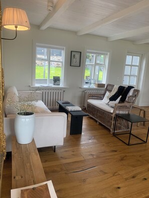 Een ruime woonkamer met een witte bank, een rieten stoel, een salontafel en een groot raam met uitzicht op het omliggende landschap.