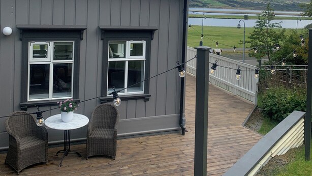 A grey building with a metal roof, two windows, a balcony with wicker chairs and a table, and a view of a lake and mountains in the background. - Aðalstræti - Historic Luxury Villa (Akureyri)