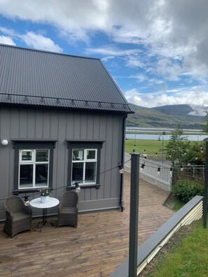 Een grijs gebouw met een metalen dak, twee ramen, een balkon met rieten stoelen en een tafel, en een uitzicht op een meer en bergen op de achtergrond.