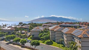 A residential area with multiple buildings, some with solar panels, a road, and a view of a mountain and ocean. - 2BR ¼ mi to Kam 1 Beach, Pool, Hot Tub, Gym, BBQ (Kihei)