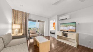 A modern living room with a sofa, a coffee table, a TV on a wooden stand, and a balcony with a view. - 2BR ¼ mi to Kam 1 Beach, Pool, Hot Tub, Gym, BBQ (Kihei)