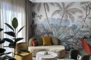 Une pièce comprenant un canapé, une table basse ronde et un fauteuil. Une grande fenêtre, ornée de rideaux, donne sur un mur décoré d’une fresque représentant un paysage tropical.