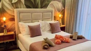 Una cama bien hecha con almohadas y una manta, una mesita de noche con una lámpara y una planta grande al fondo.