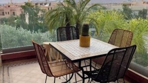 Un balcón con mesa y sillas, una planta en maceta y vistas a edificios y follaje.