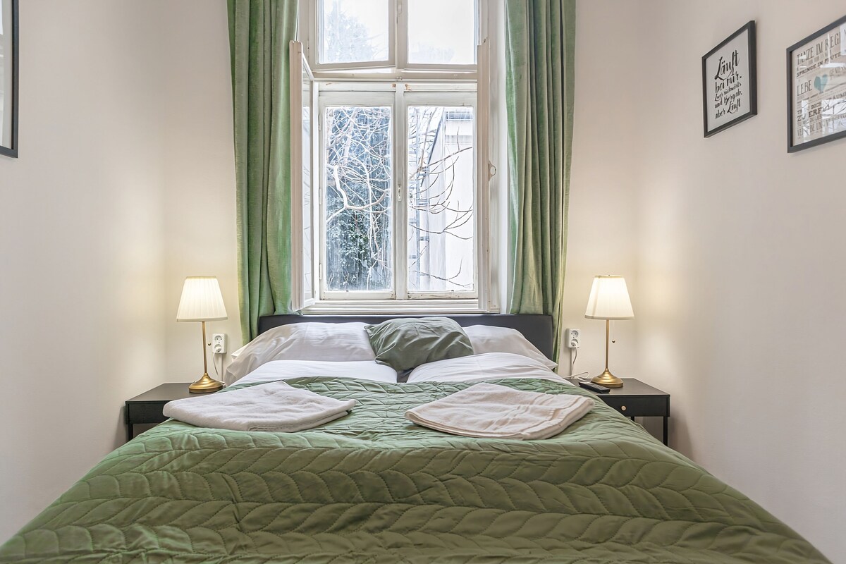 A neatly made bed with a green quilt, two bedside lamps, and a window with curtains.