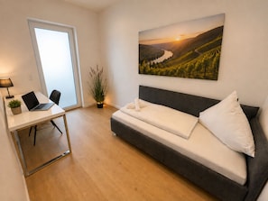 Een kamer met een bed, een bureau met een laptop, een kamerplant en een schilderij van een landschap.