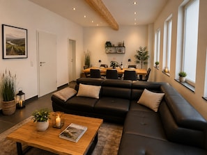 Een moderne woonkamer met een zwarte leren bank, een houten salontafel met een tijdschrift, en een eethoek op de achtergrond.