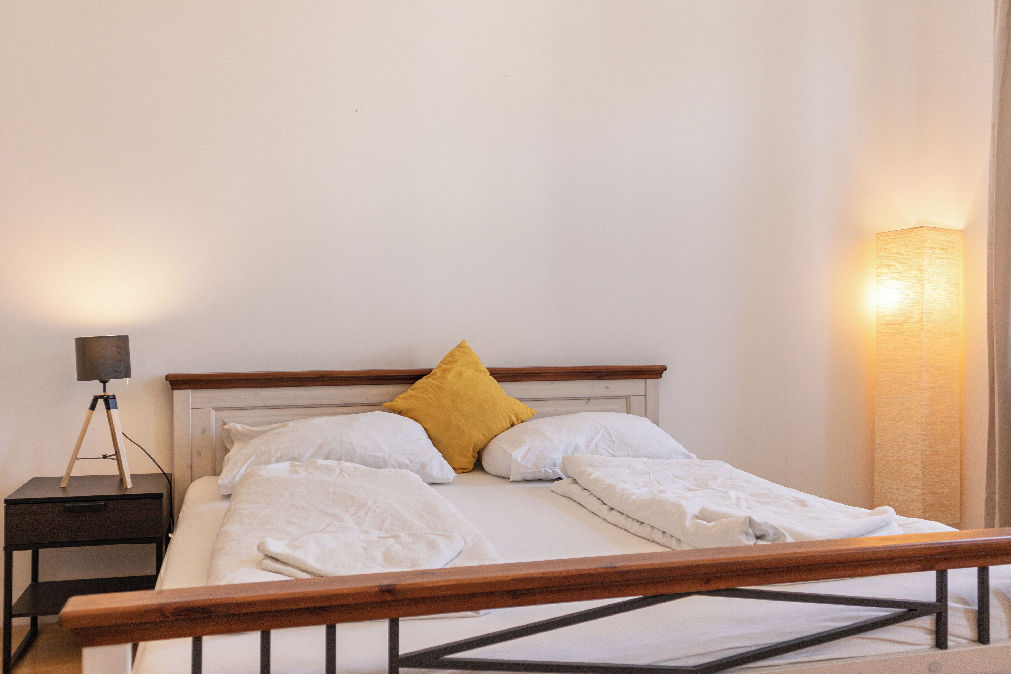 Un letto rifatto con lenzuola bianche e un cuscino giallo senape, un comodino con una lampada e un punto luce a parete.