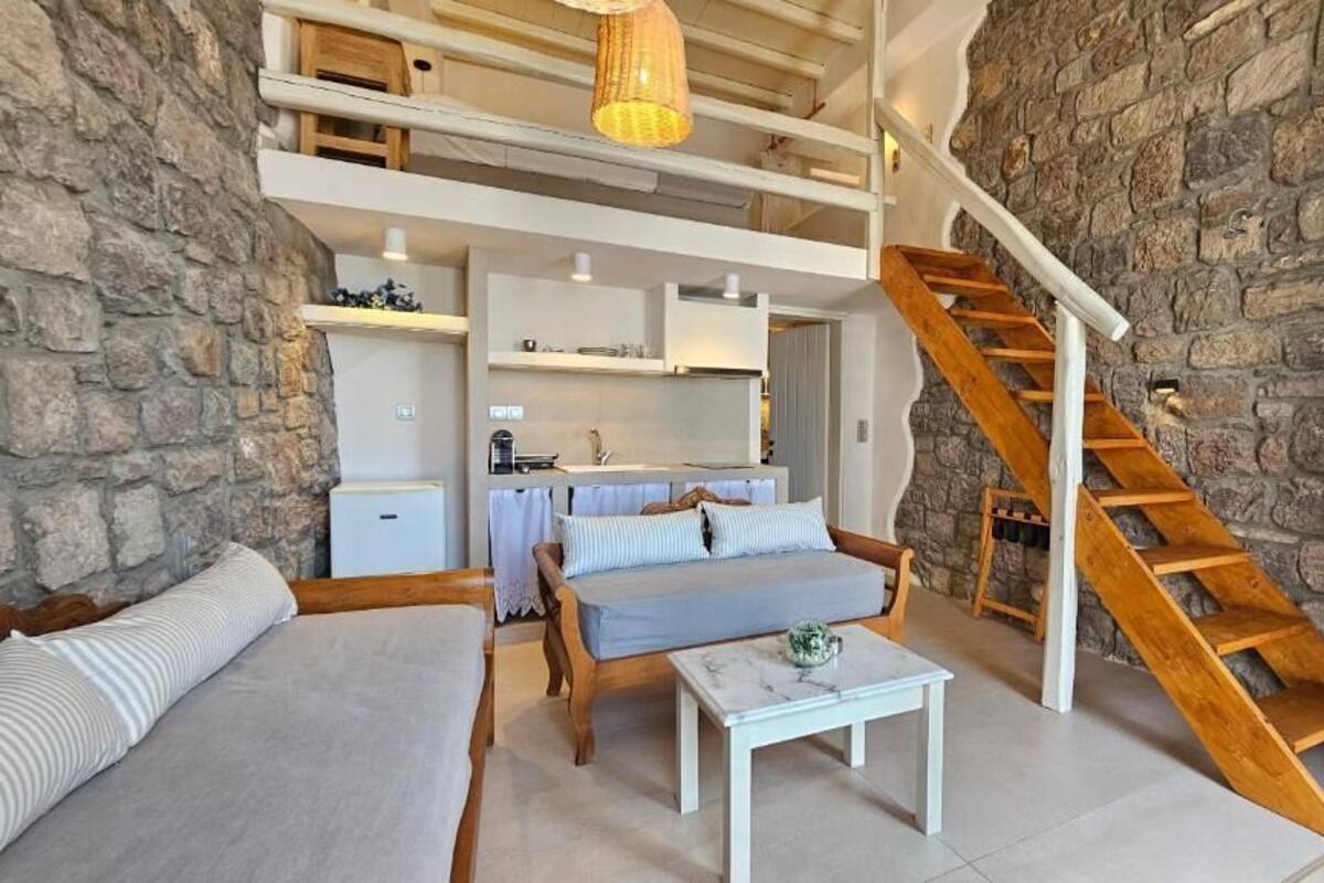 Un espacio habitable tipo loft con una pared de piedra, una escalera de madera y una cocina visible a través de una puerta abierta.