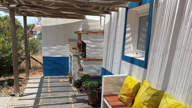 A covered outdoor area with a stone-tiled floor, a wooden bench with yellow cushions, and a white wall with a window.