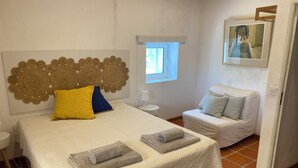 A bedroom with a bed, a bedside table, a chair, a window, and a painting on the wall.