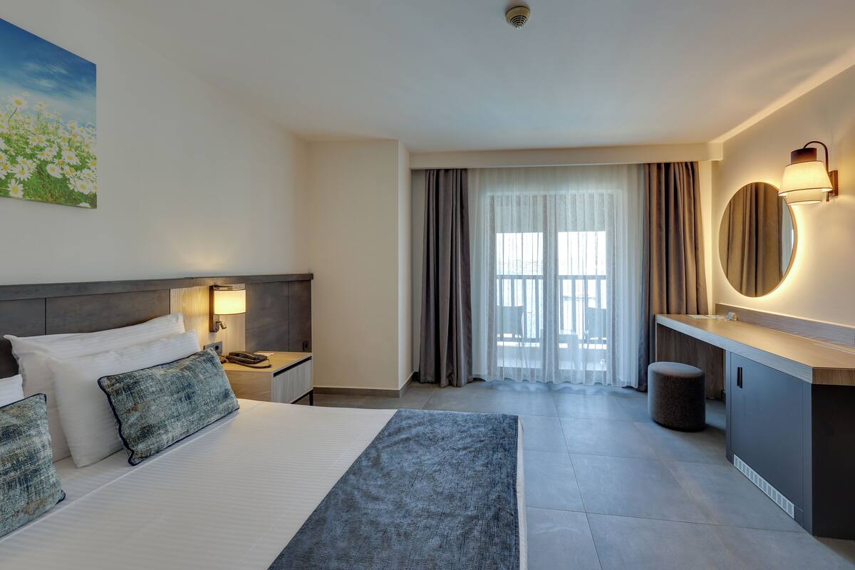 Ein modernes Hotelzimmer mit einem großen Bett, Nachttischlampen, einem runden Spiegel, einem kleinen runden Hocker und Blick ins Freie durch ein Fenster mit Vorhängen.