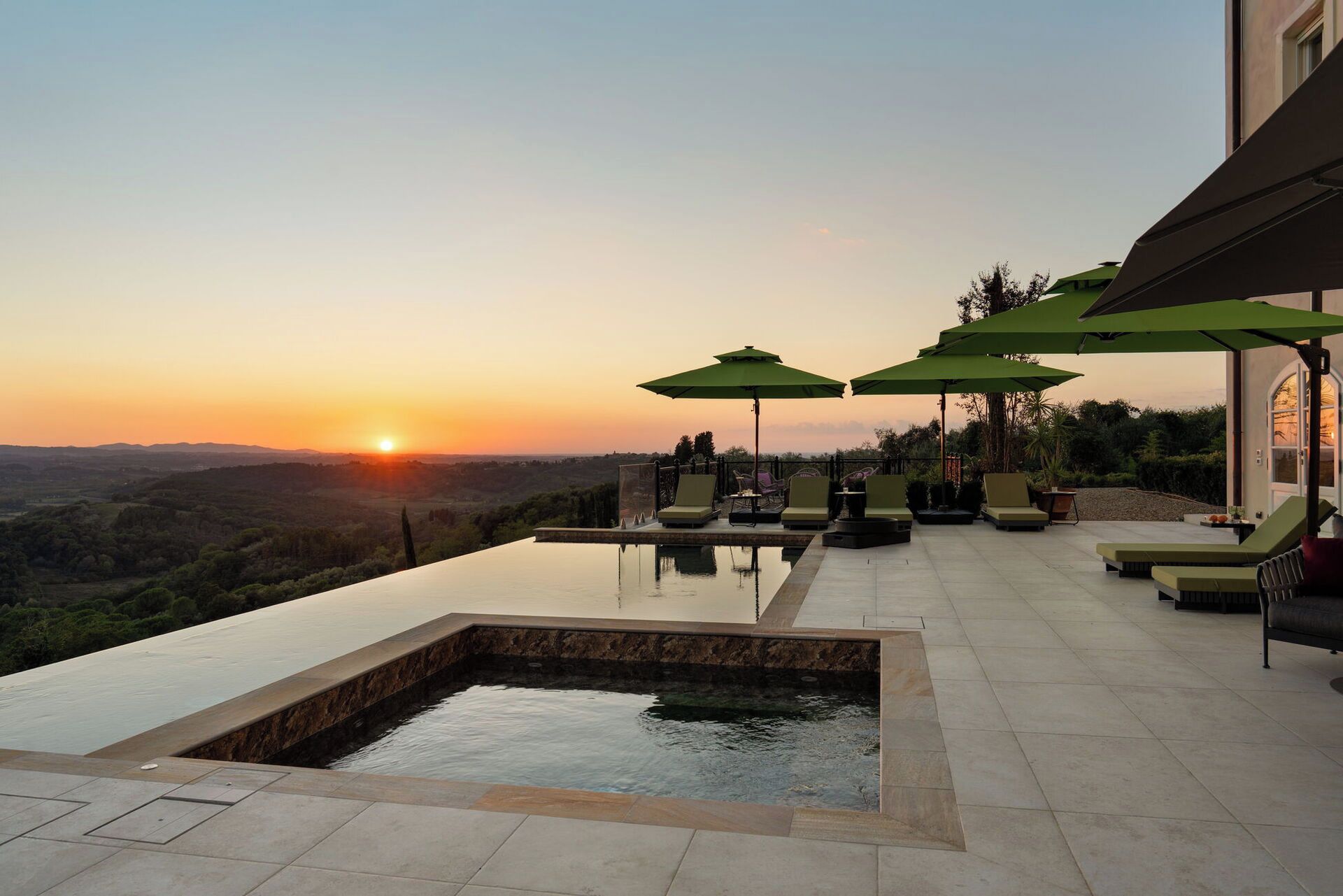 An infinity pool with a view of the sunset, outdoor seating with umbrellas, and a landscaped garden.