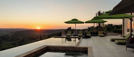 An infinity pool with a view of the sunset, outdoor seating with umbrellas, and a landscaped garden.