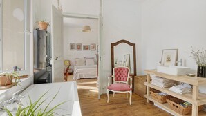 A room with a wooden floor, a white bathtub, a wooden chair, a shelf with towels, and a door leading to another room.