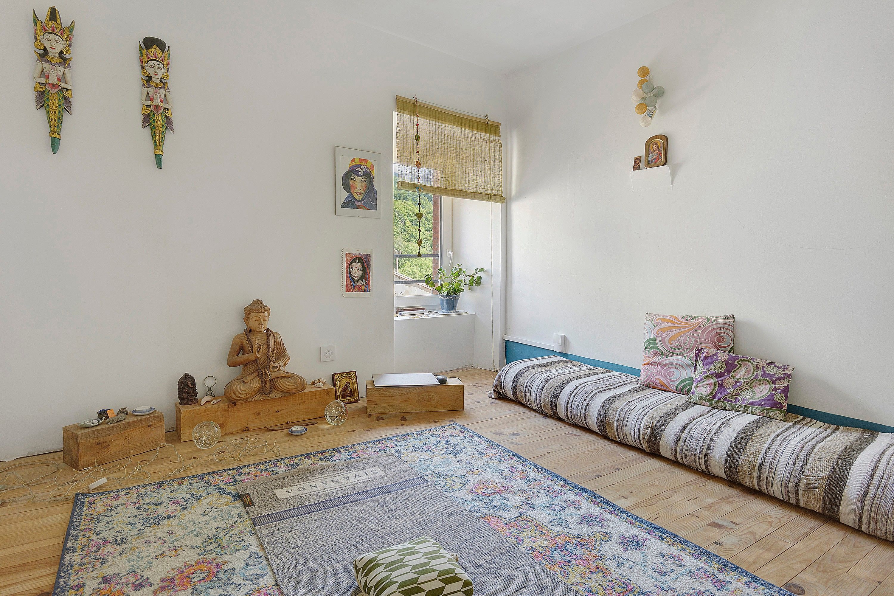 Una habitación con piso de madera, un tapete estampado, una cama con cabecera azul, un armario de madera y decoraciones en la pared, incluyendo una estatua de Buda y fotografías enmarcadas.
