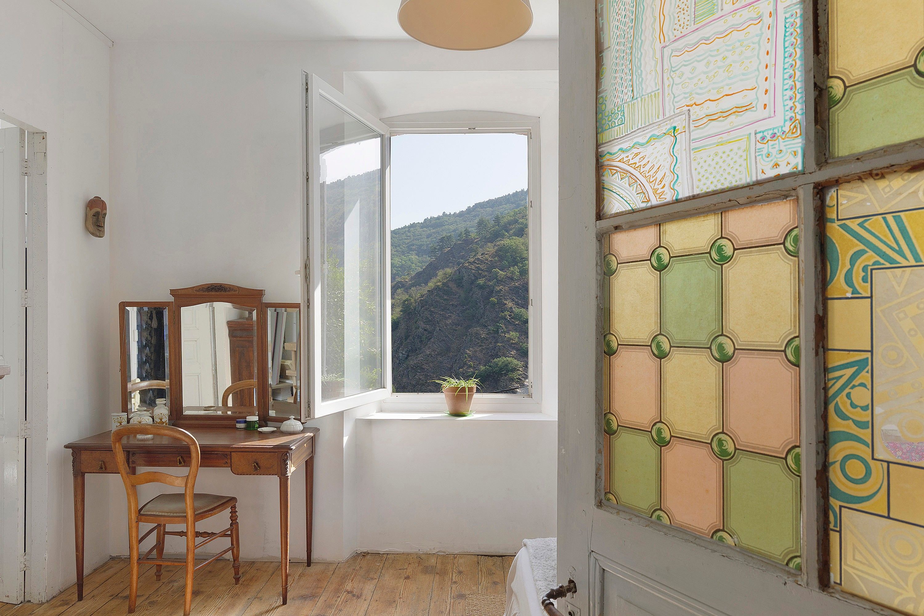 Quarto com escrivaninha de madeira, uma cadeira, janela com vista para uma montanha e um painel decorativo na parede.