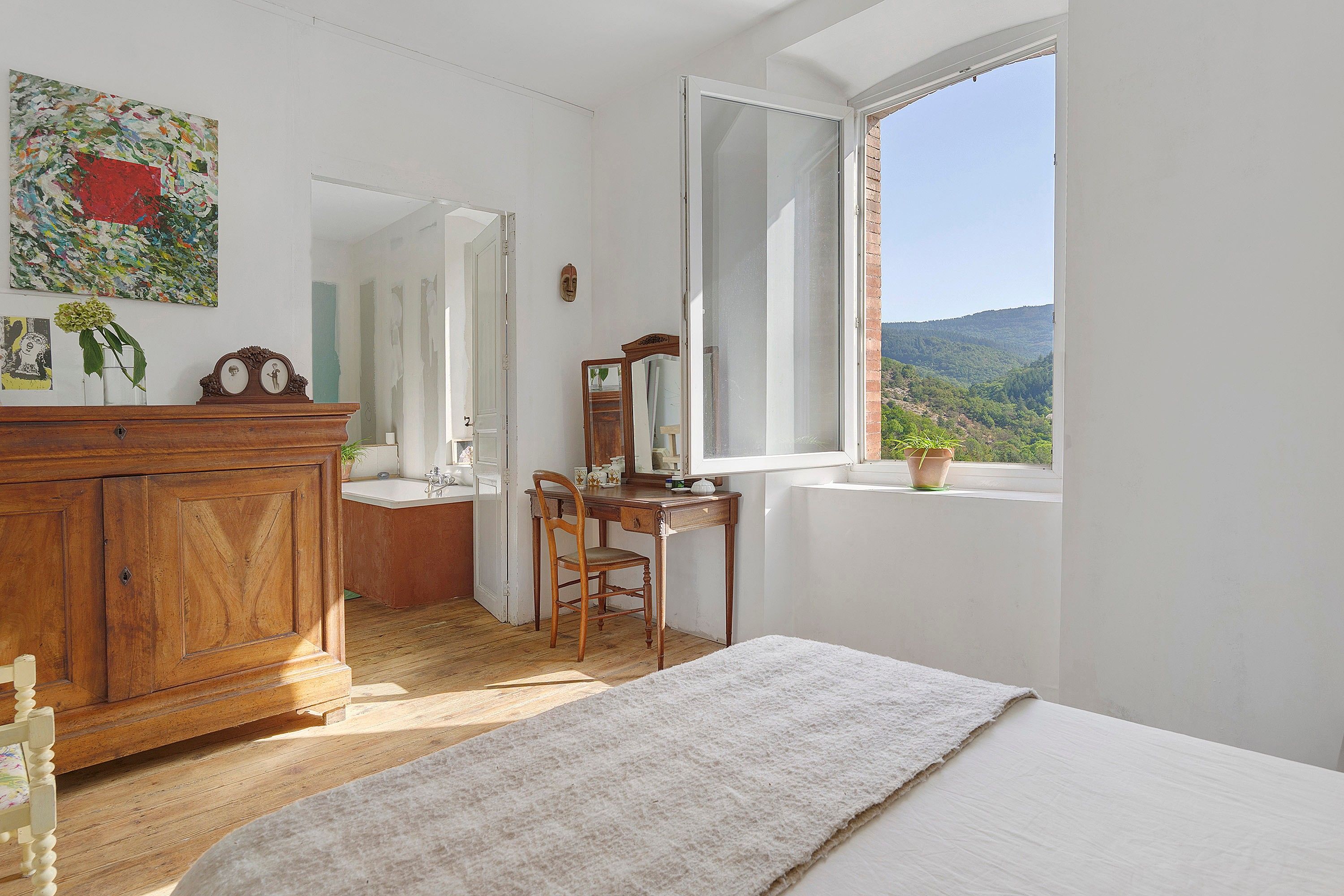 A bedroom with a wooden dresser, a desk with a chair, a large window with a view of the outdoors, and a painting on the wall.