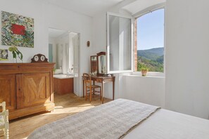 Ein Schlafzimmer mit einem Holzkoffer, einem Schreibtisch mit Stuhl, einem großen Fenster mit Blick ins Freie und einem Bild an der Wand.