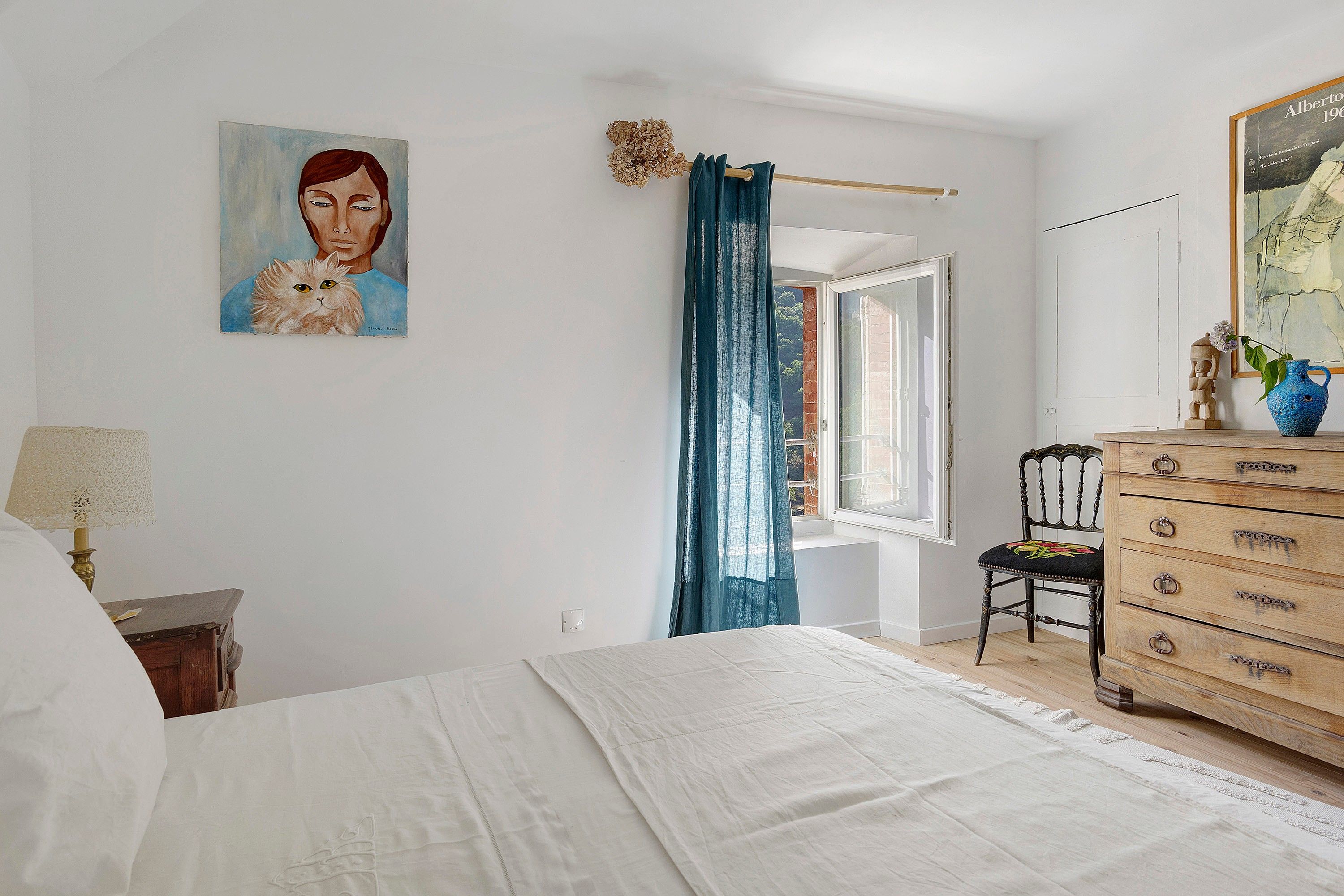 A bedroom with a bed, a dresser, a chair, a painting, and a window with curtains.