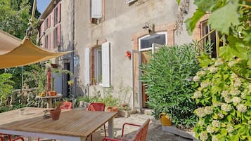 A courtyard with a wooden table and chairs, surrounded by potted plants and a building with a window and shutters.