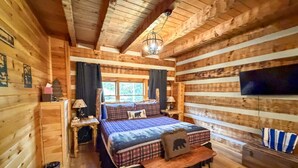 Ein gemütliches Schlafzimmer in einer Blockhütte mit einem Bett, einem Nachttisch, einer Lampe und einem an der Wand befestigten Fernseher.