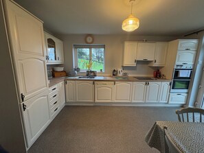 A kitchen with white cabinetry, a window with a view of greenery, a clock, and a chandelier. - Jerry's Cottage, Dingle Penninsula Wild Atlantic Way and Páirc Nasiúnta na Mara (Co)