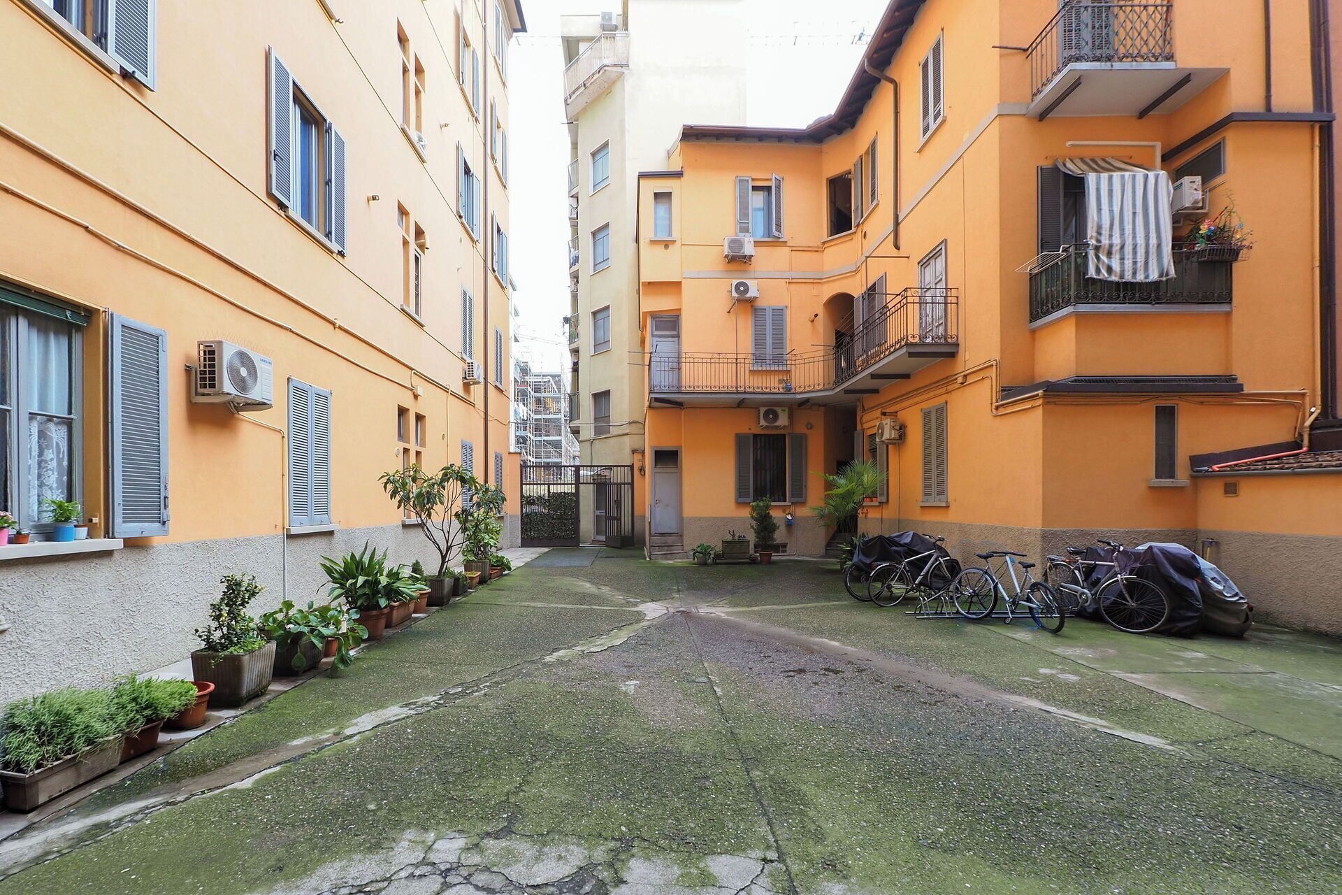 A narrow alleyway between two yellow buildings with balconies, potted plants, and bicycles parked along the sides.