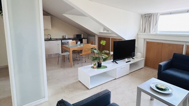 A modern living room with a flat-screen TV, a dining area with a table and chairs, and a kitchen in the background. - Attic Gardens Flexrent - Abyssinia (Riccione)