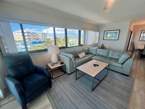 A modern living room with a sectional sofa, a coffee table, and a blue armchair. There is a large window with a view of buildings and a lamp on a side table. - Best view on Siesta Key | Walk Right onto Siesta Key Beach (Sarasota)