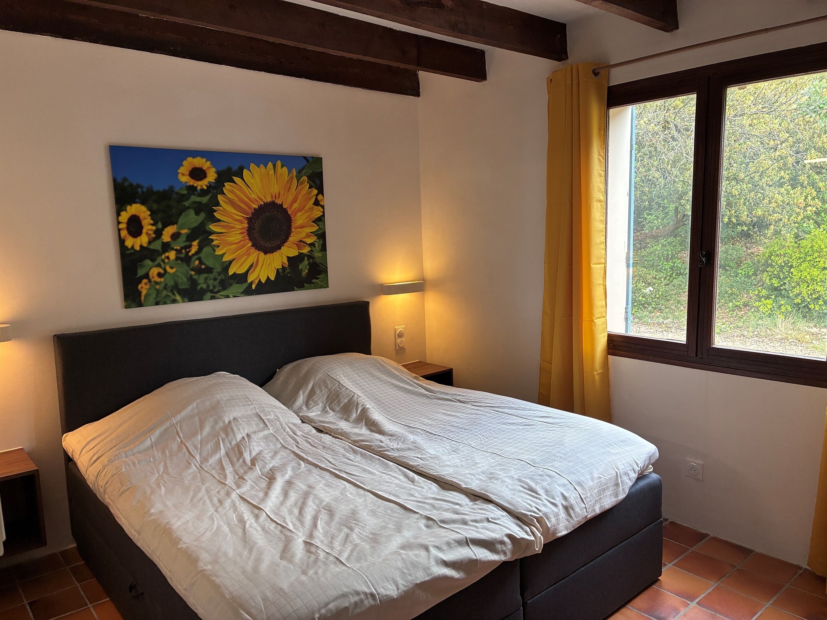 Ein Bett mit Kopfteil, ein Wandbild mit Sonnenblumen, eine Nachttischlampe und ein Fenster mit Vorhängen.