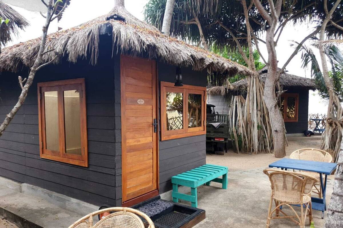 A small wooden cabin with a thatched roof, a wooden door, and a window. There is a bench and a table with chairs outside.