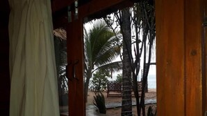 Blick aus einem Zimmer mit Holztür und Fenster auf einen Strand mit Palmen und einer Holzbank.