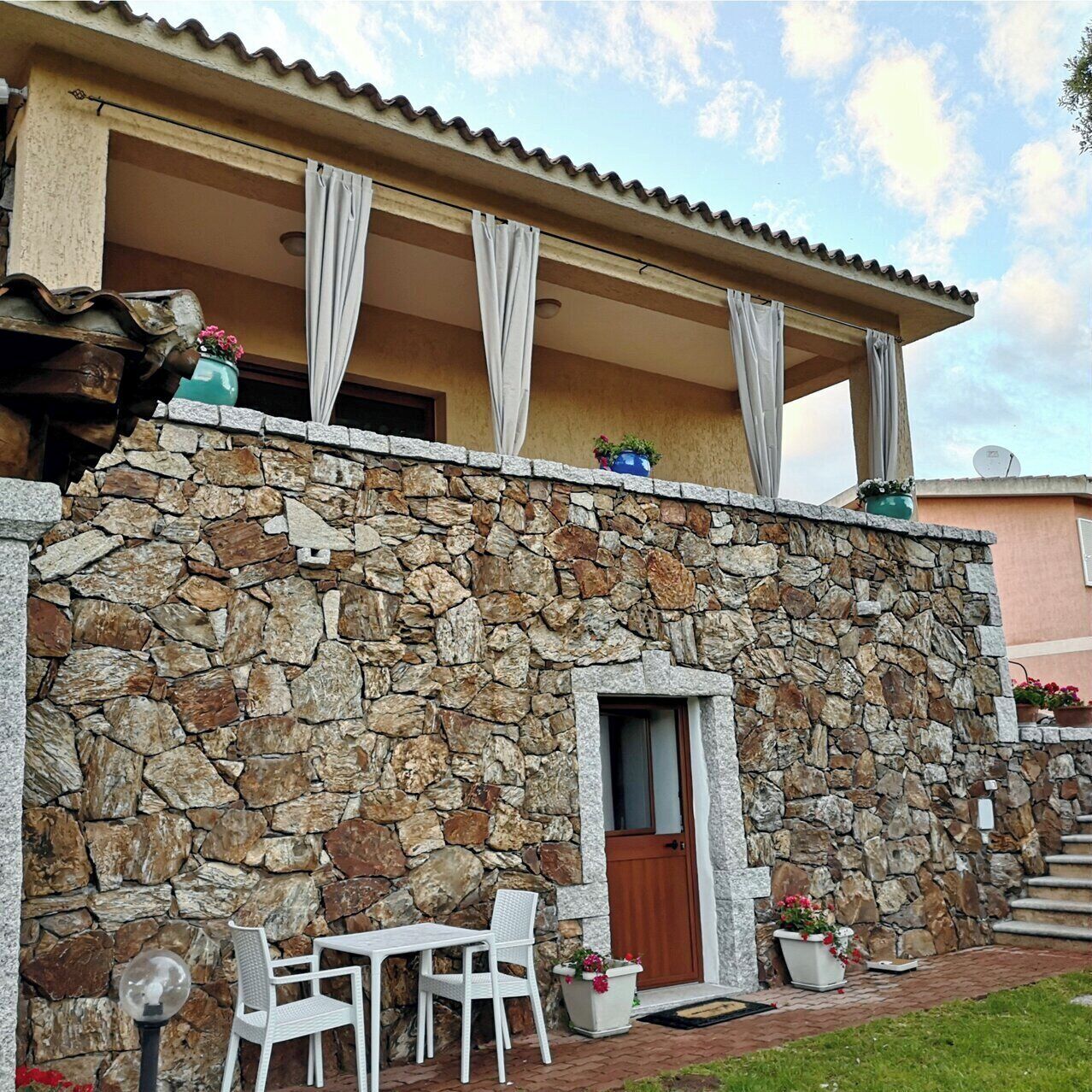 Fachada de pared de piedra con una puerta, un pequeño espacio al aire libre con mesa y sillas, y plantas en macetas.