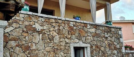 Fachada de pared de piedra con una puerta, un pequeño espacio al aire libre con mesa y sillas, y plantas en macetas.