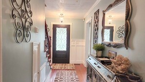 Un pasillo con alfombra estampada, una puerta oscura con un panel de vidrio decorativo, una escalera de madera y una consola con objetos decorativos.