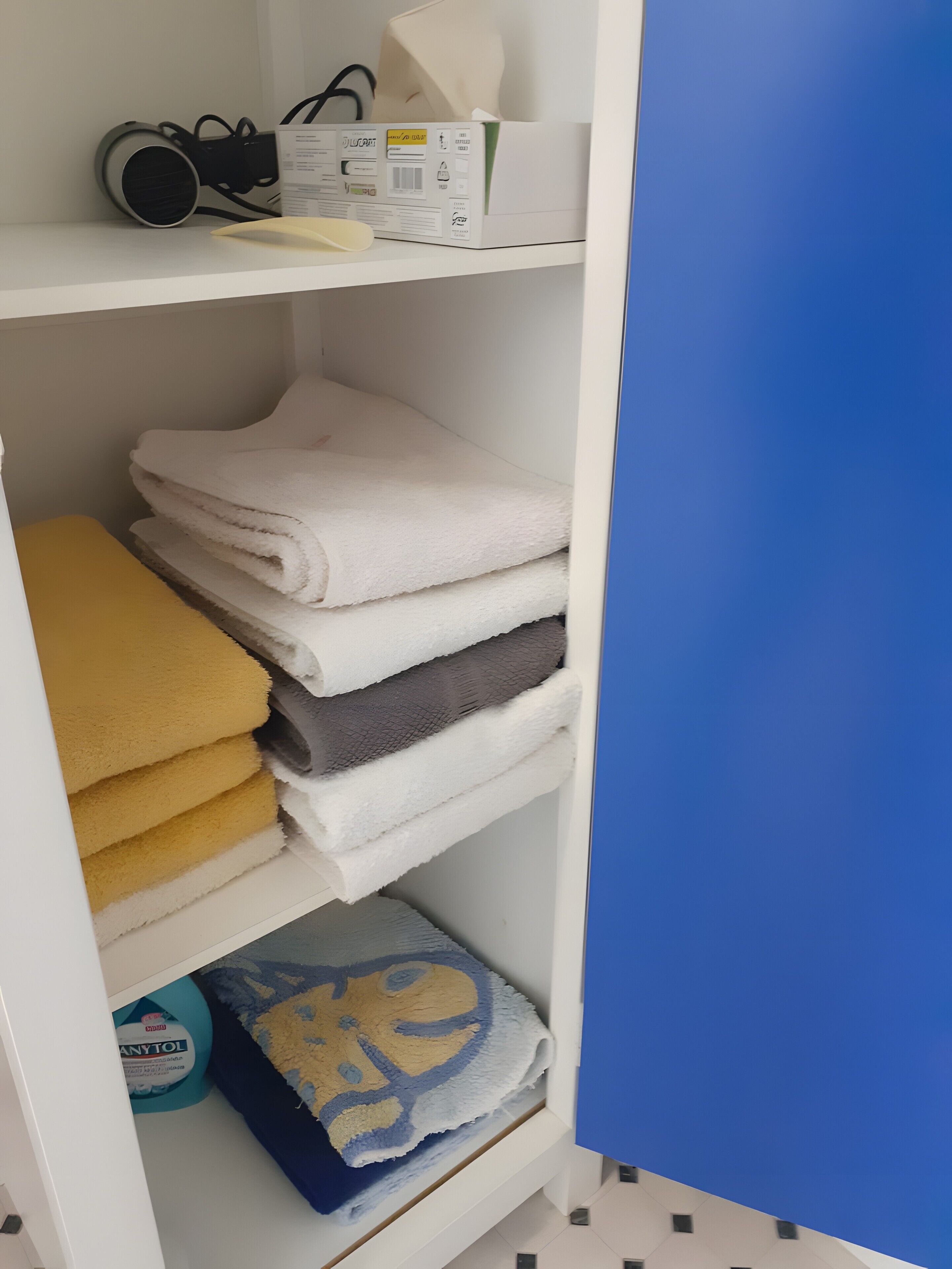 Un estante blanco con toallas dobladas en blanco, amarillo y gris, una caja con una toalla blanca y una pared azul.